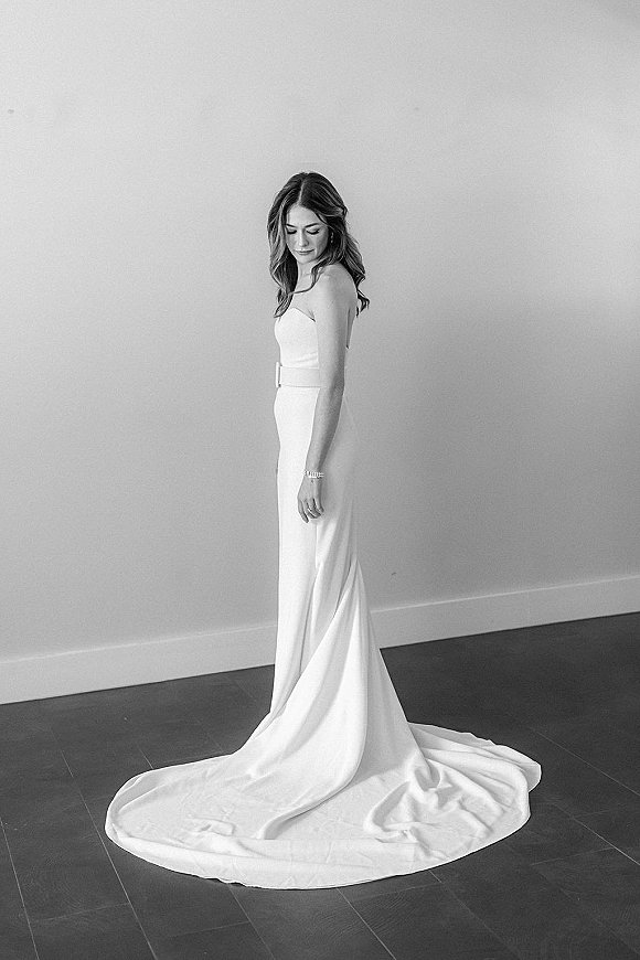 Bridal portrait in black and white of a bride looking down in a strapless wedding dress with sash belt and long train against a plain wall
