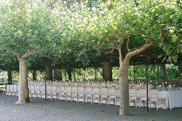 Outdoor reception tablescape with a long banquet table wedding setup, white linens, floral centerpieces, candles, and string lights under trees