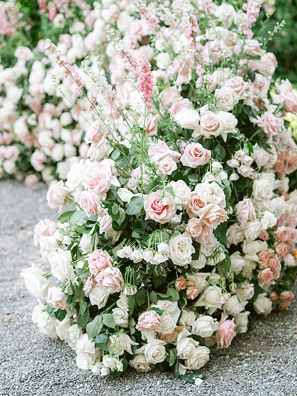 Wedding floral arrangement of pink and white roses with greenery accents on gravel ground, set against a softly blurred floral backdrop
