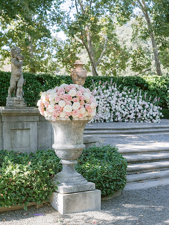 Wedding floral arrangement in a stone urn with blush and white roses and greenery on stone steps beside a garden statue on a terrace