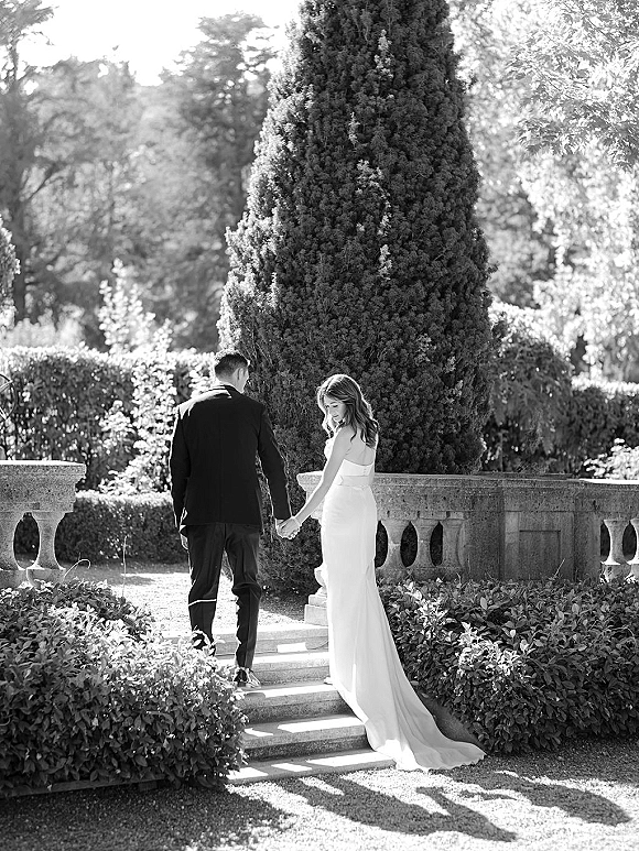 Couple portrait of bride and groom holding hands, walking up garden stone steps, bride looking back in strapless dress with train