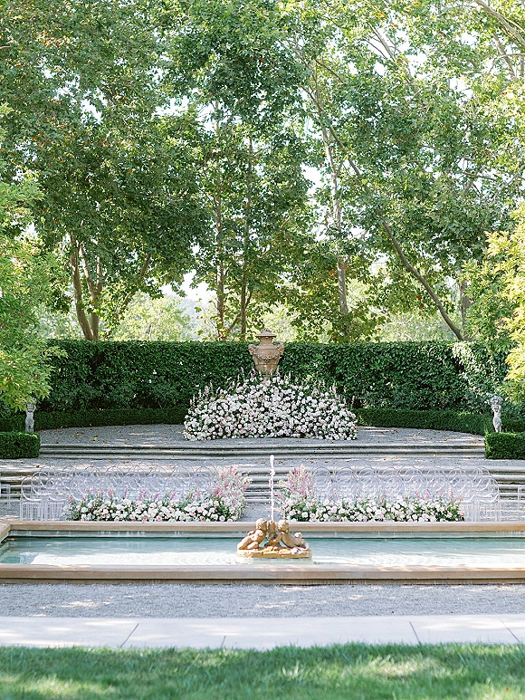 Outdoor ceremony decor with clear acrylic chairs flanking floral-lined stone steps, framed by a fountain, urns, statues, and boxwood hedges