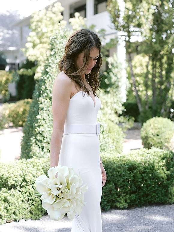 Bridal portrait of a bride in a strapless wedding dress holding a white calla lily bouquet, looking down on a gravel garden path.