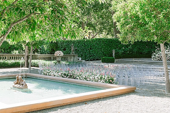 Outdoor ceremony setup with clear acrylic ceremony chairs lining a floral aisle border of pink and white flowers beside a fountain pool courtyard