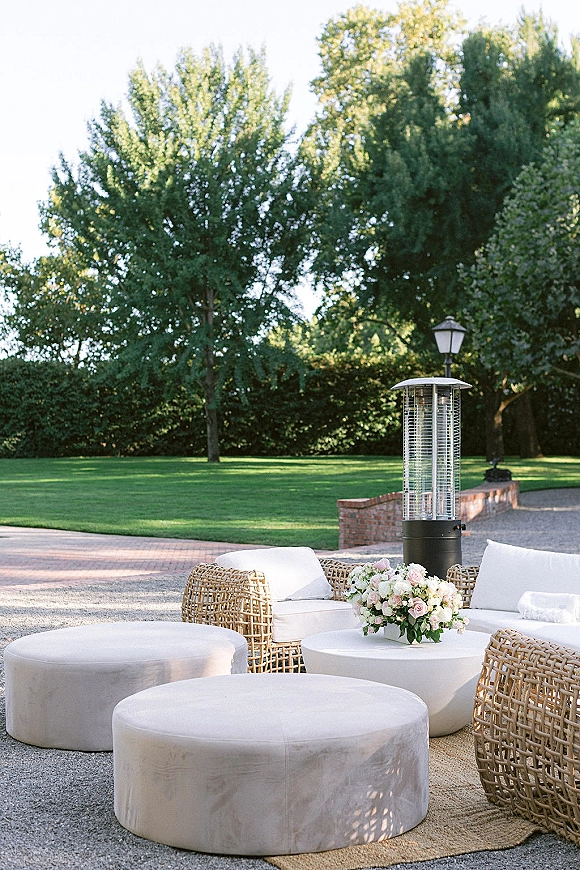 Outdoor lounge seating with wicker sofas, white cushions, and round ottomans around a low table with floral centerpiece on a gravel patio lawn