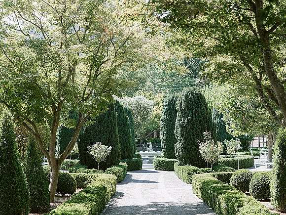 Garden aisle outdoor ceremony aisle with a gravel path, white chairs, trimmed hedges and topiary, leading to a fountain in sunlight
