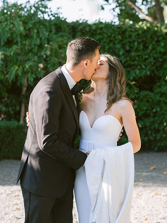 Wedding kiss portrait of bride and groom kissing in side profile, her strapless dress and his black tuxedo against a green hedge walkway