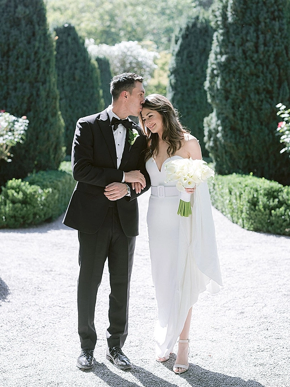 Couple portrait of groom kissing bride’s forehead as she holds a white calla lily bouquet, arm in arm on a sunlit garden gravel path