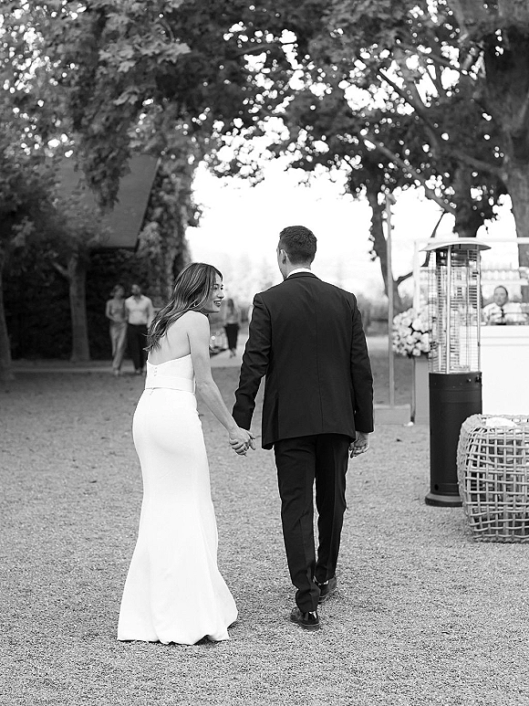 Couple portrait of newlyweds holding hands walking away as the bride looks back, strapless gown and groom suit on a tree-lined gravel path with guests behind