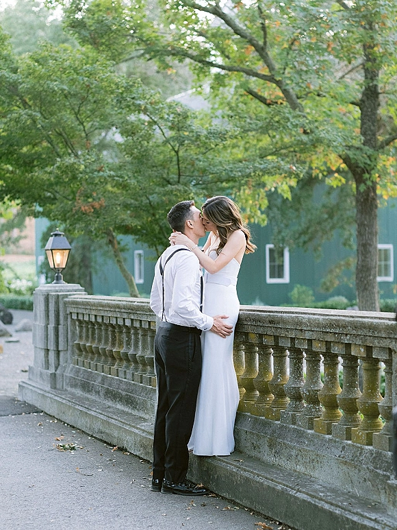 Wedding kiss portrait of bride and groom kissing on a stone bridge, her strapless dress train flowing as he wears suspenders near trees and barn