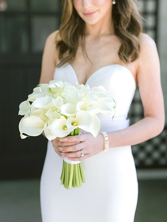Bridal portrait of bride holding bouquet, a white calla lily bridal bouquet over a strapless gown in soft light by a dark doorway
