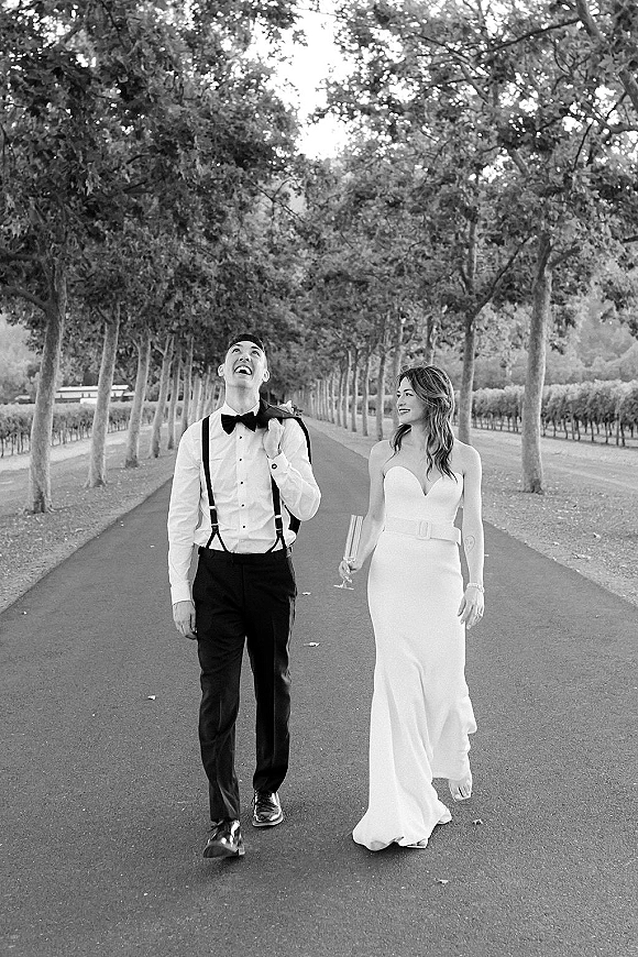 Couple portrait of bride and groom walking, laughing on a tree-lined vineyard road, she holds a champagne flute, he wears suspenders and bow tie