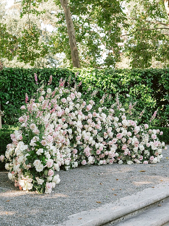 Wedding floral arrangement of pink and white roses with greenery and tall pink stems lining a gravel garden path by a hedge