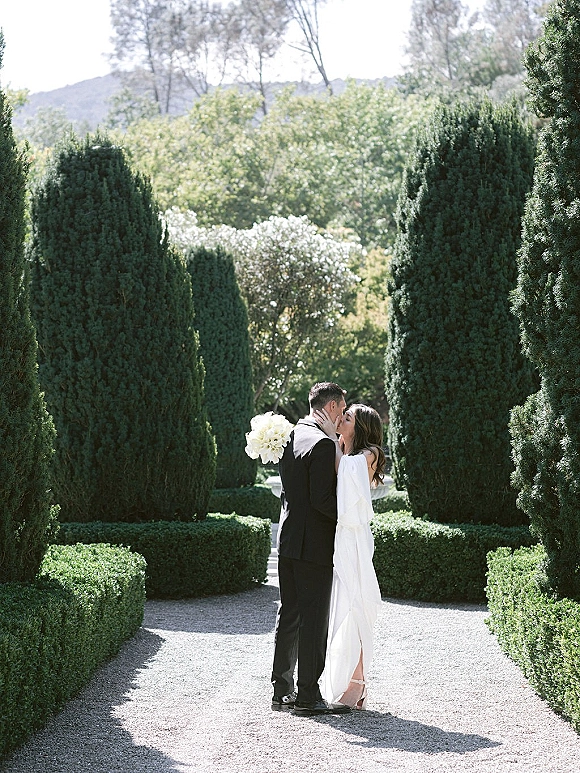 Wedding kiss as bride and groom kiss on a gravel path in a formal garden, bride holding a white bouquet in an off-shoulder gown