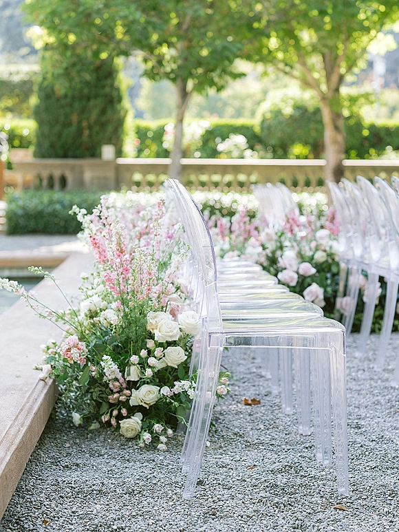 Ceremony aisle decor with white roses and blush flowers lining a gravel runner, flanked by clear acrylic chairs on a garden terrace