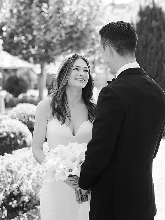 Couple portrait in a black and white wedding portrait style, bride in strapless dress holding calla lily bouquet as groom in tux gazes at her in a garden bokeh setting