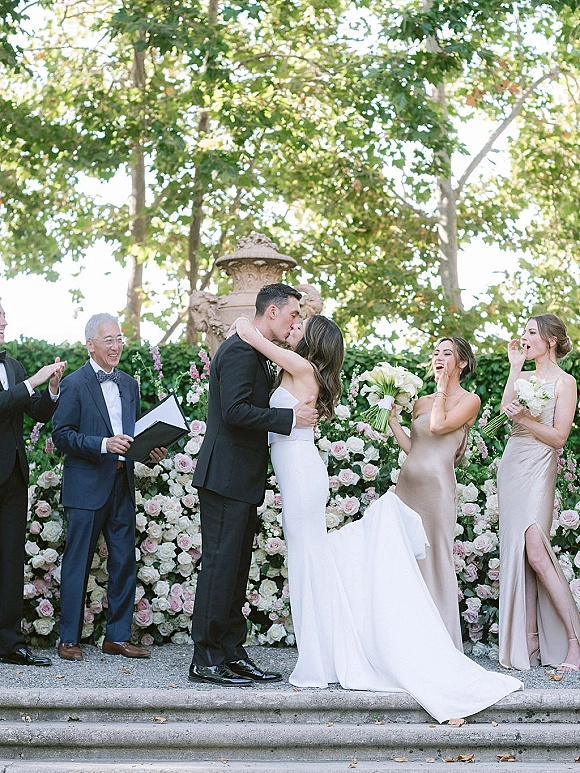Ceremony kiss as bride in strapless gown and groom in tux embrace at outdoor altar, bridal party cheering by floral wall and stone steps