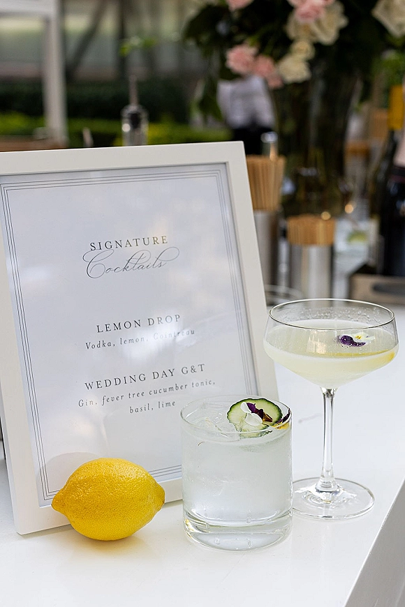 Signature cocktails sign in a framed bar menu display beside cocktail glasses with lemon, ice, cucumber garnish, and edible flowers outdoors