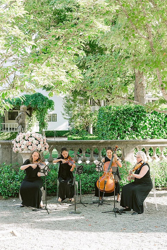 Wedding string quartet in black dresses playing on clear chairs with pink rose accent, music stands and mics in a garden courtyard