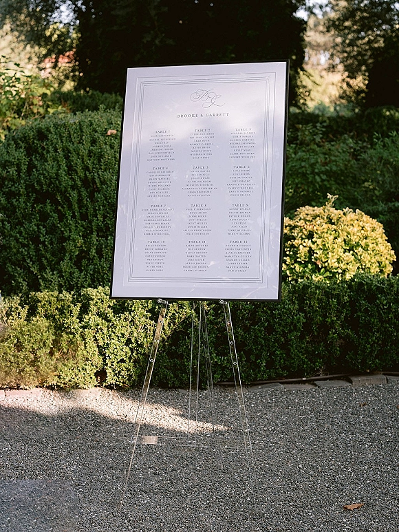 Wedding seating chart with calligraphy text in a framed sign on a clear acrylic easel, set on a gravel path amid garden greenery