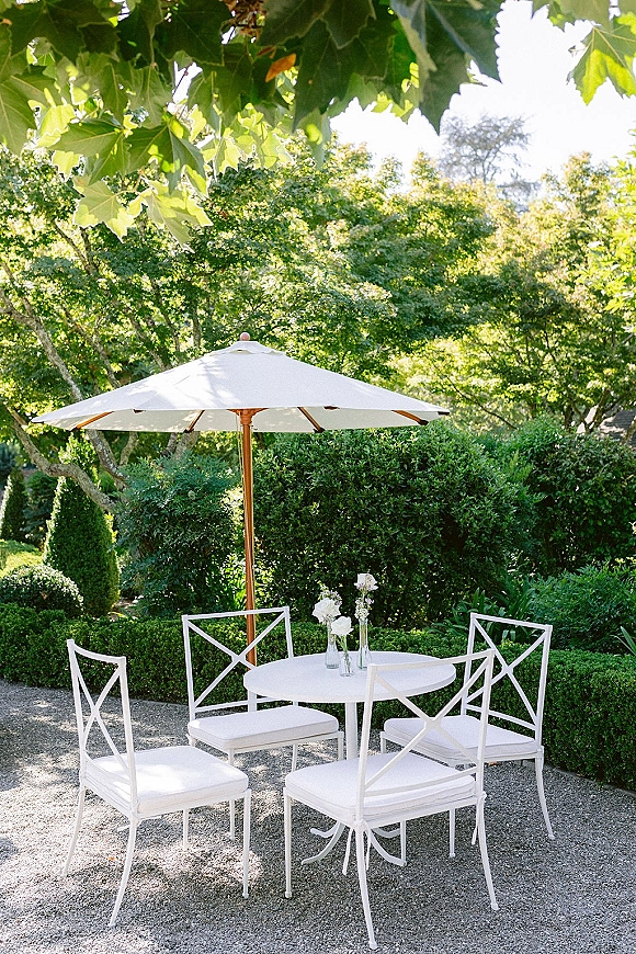 Outdoor cocktail seating with a patio umbrella over a round table, white metal chairs with cushions, and white bud vases on a gravel patio amid hedges