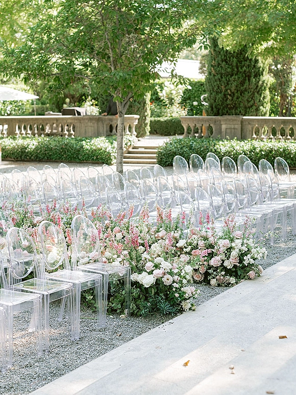 Ceremony aisle decor with acrylic wedding chairs lining blush and white rose florals along a gravel path beside stone steps and trees