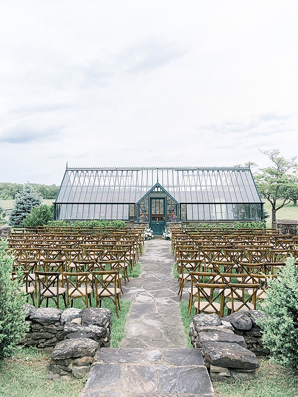 Ceremony setup with outdoor wedding ceremony chairs lined along a stone aisle, white florals and greenery leading to a greenhouse backdrop