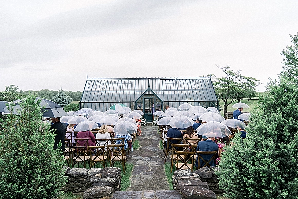 Outdoor ceremony setup with clear umbrellas over crossback chairs lining a stone aisle, white florals and greenery by a greenhouse under gray skies