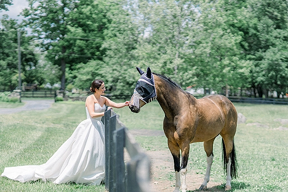 Bridal portrait of a bride with horse in a strapless full-skirt gown and long train by a wooden fence, with a green pasture behind