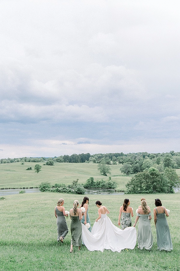 Bridesmaids with bride walking away across a grassy field, carrying her dress train and bouquets under a cloudy sky with hills and pond