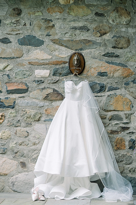 Wedding dress hanging on a wall hook, strapless wedding dress with tulle veil draped over it, bridal shoes below against stone wall