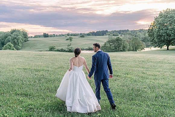 Couple portrait of bride and groom walking hand in hand in a grassy field at sunset, her full skirt and his suit with pond beyond