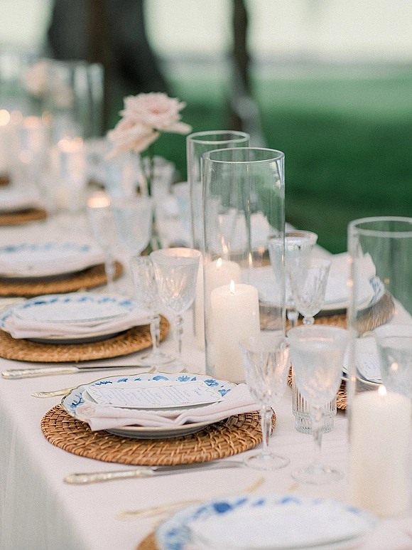 Reception tablescape wedding table setting with rattan placemats, patterned plates, crystal glassware, and candlelit hurricane vases on a lawn backdrop