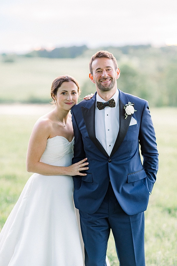 Couple portrait of bride and groom smiling, bride holding his arm in a strapless gown and navy tuxedo in a green hill field