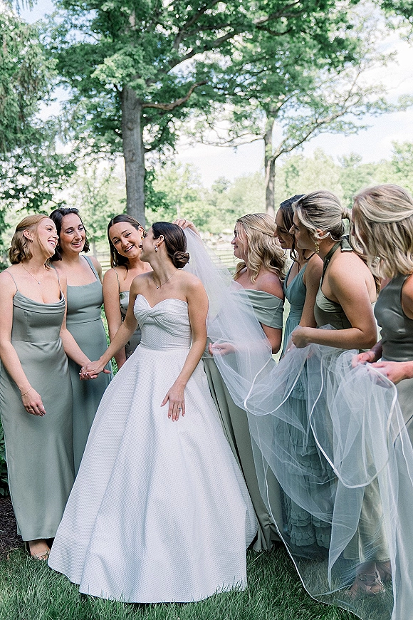 Bride with bridesmaids laughing in sage green dresses, holding her long veil over a grassy park lawn with trees in daylight