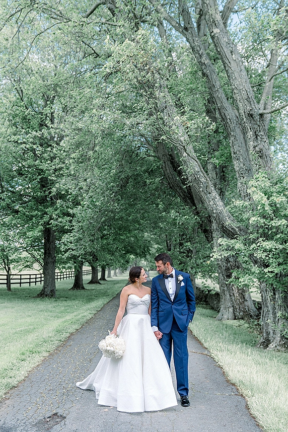 Couple portrait of bride and groom walking hand in hand, her strapless ballgown and white bouquet on a tree-lined road