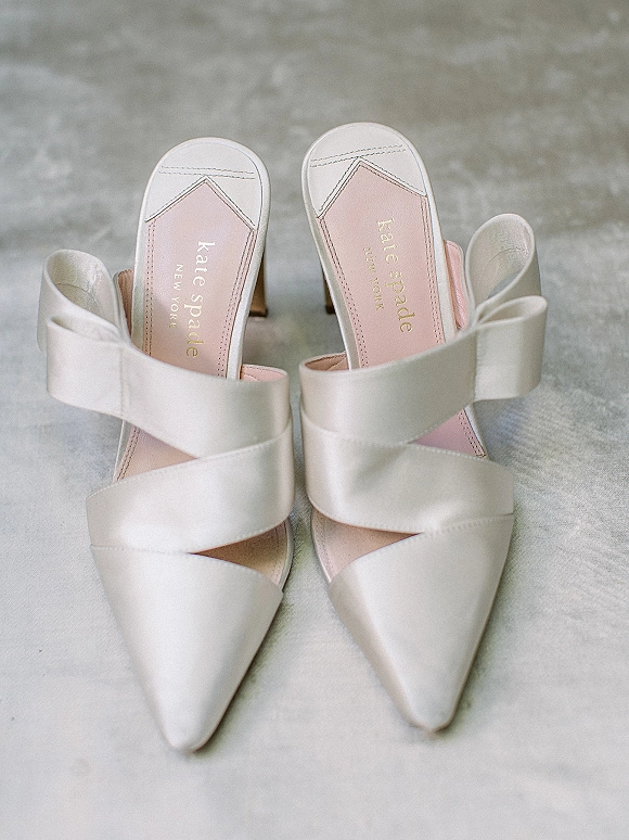 Bridal shoes in white satin mules with pointed toes, criss cross straps, and block heels on a light gray textured surface