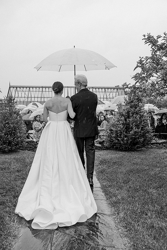 Wedding processional with bride walking down aisle under a white umbrella beside a suited escort on a stone path as guests hold umbrellas in light rain