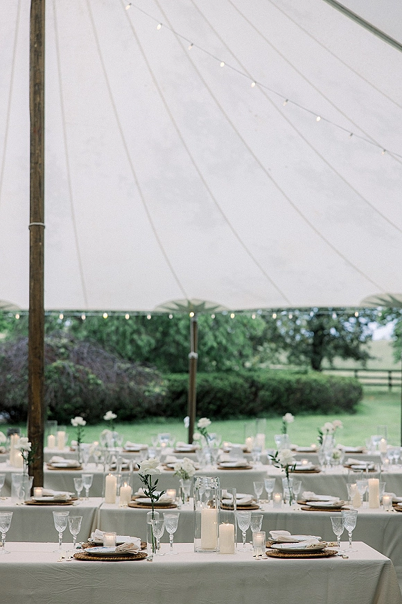 Reception tablescape with white linens, taper candles in glass hurricanes, crystal stemware and woven chargers under a sailcloth tent with string lights