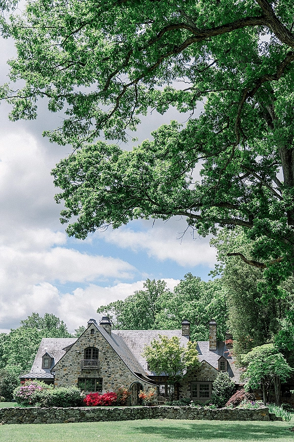 Wedding venue exterior with a stone cottage and slate roof, framed by flowering shrubs and a stone wall under a blue sky