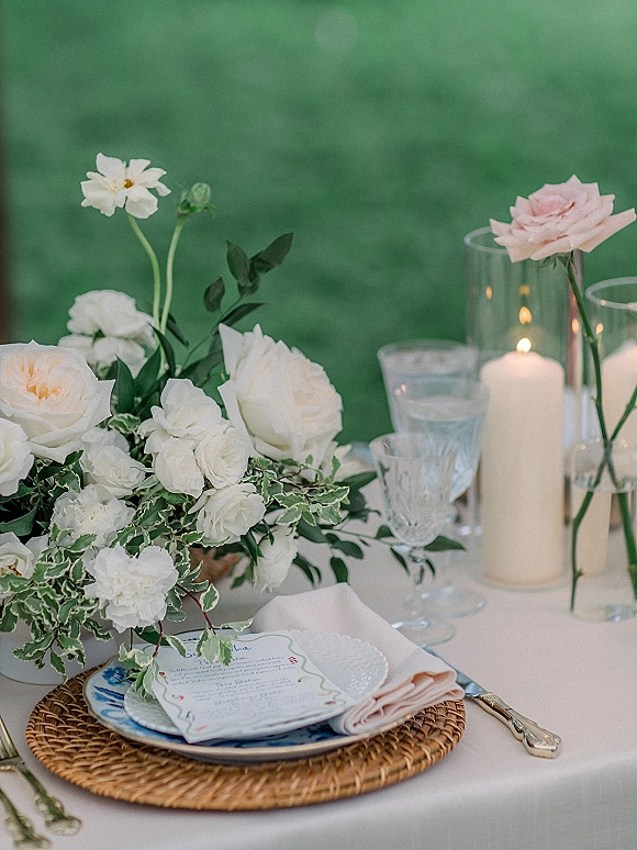 Reception tablescape with wedding table centerpiece of white roses and greenery, taper candles and rattan chargers set on a green lawn