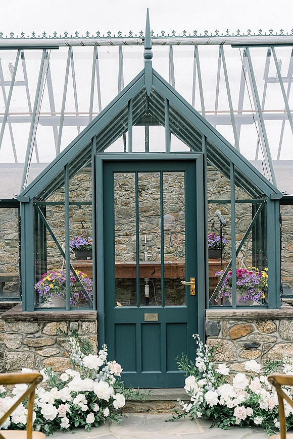 Wedding ceremony backdrop with white and blush roses and greenery garland framing glass greenhouse doors on a stone patio