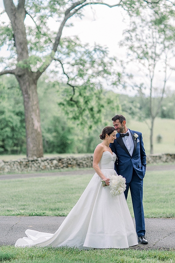 Couple portrait of an outdoor bride and groom smiling, her strapless dress and white bouquet beside his navy tuxedo on a path by trees and stone wall