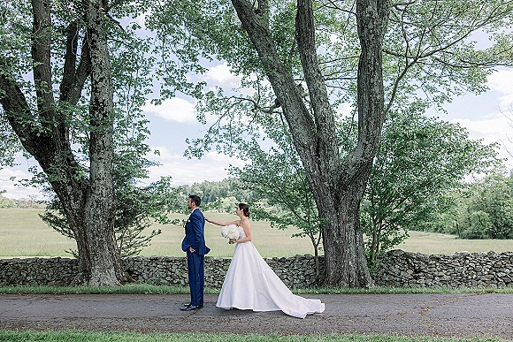 Wedding first look as bride taps groom’s shoulder, holding a white bouquet beside a stone wall under large trees and a cloudy sky