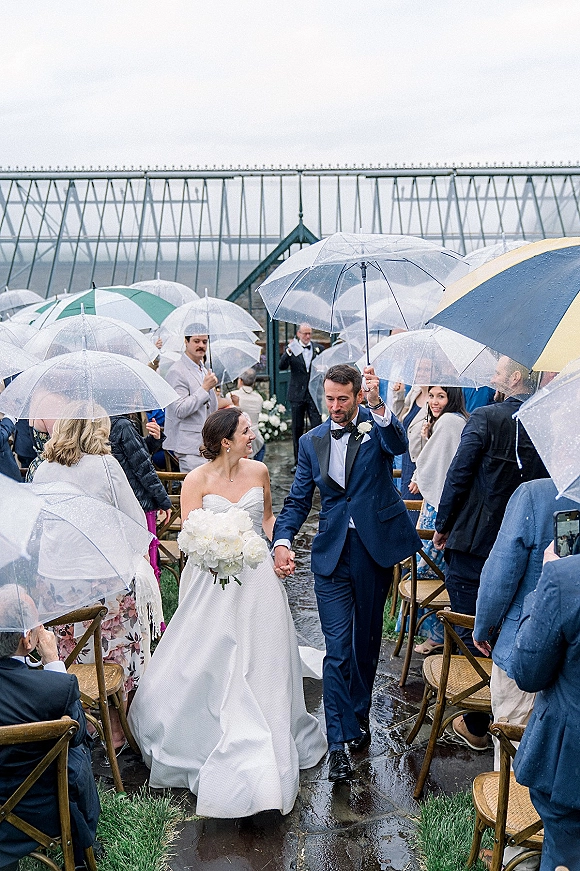 Wedding recessional as bride and groom walking aisle under a clear umbrella on wet pavement, guests lining the outdoor aisle by a greenhouse