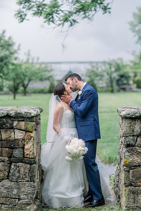 Wedding kiss portrait of bride and groom kissing, her veil flowing and white bouquet held close, by a stone wall on an overcast lawn