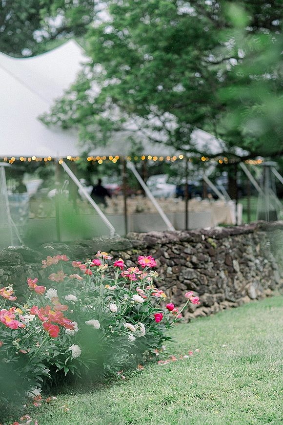 Outdoor reception tent with clear top, glowing string lights, and round white linen tables beside a floral garden border and stone wall