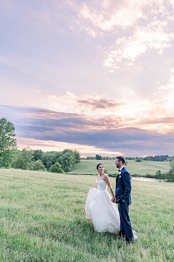 Couple portrait of bride and groom outdoors holding hands, she lifts her dress and looks at him in a grassy field at sunset.
