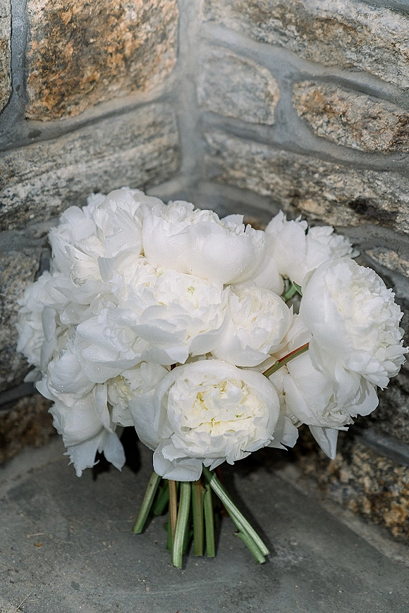 Bridal bouquet of white peonies with visible stems resting on a stone floor against a stone wall backdrop