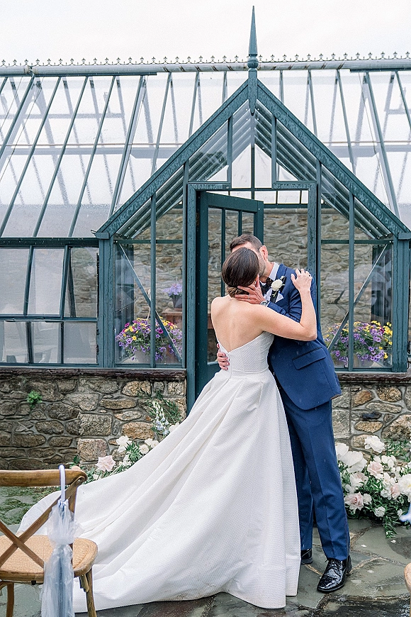 Wedding kiss portrait of bride and groom kissing, her strapless gown with long train beside a glass greenhouse on a stone patio
